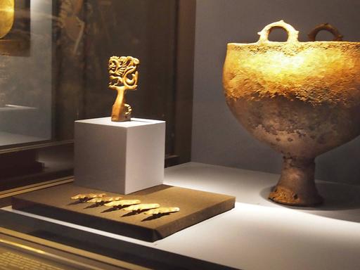 Ein großer goldfarbener Kessel, fünf Kleiderspangen und andere Kunstobjekte sind in einer Vitrine in einem Museum ausgestellt.