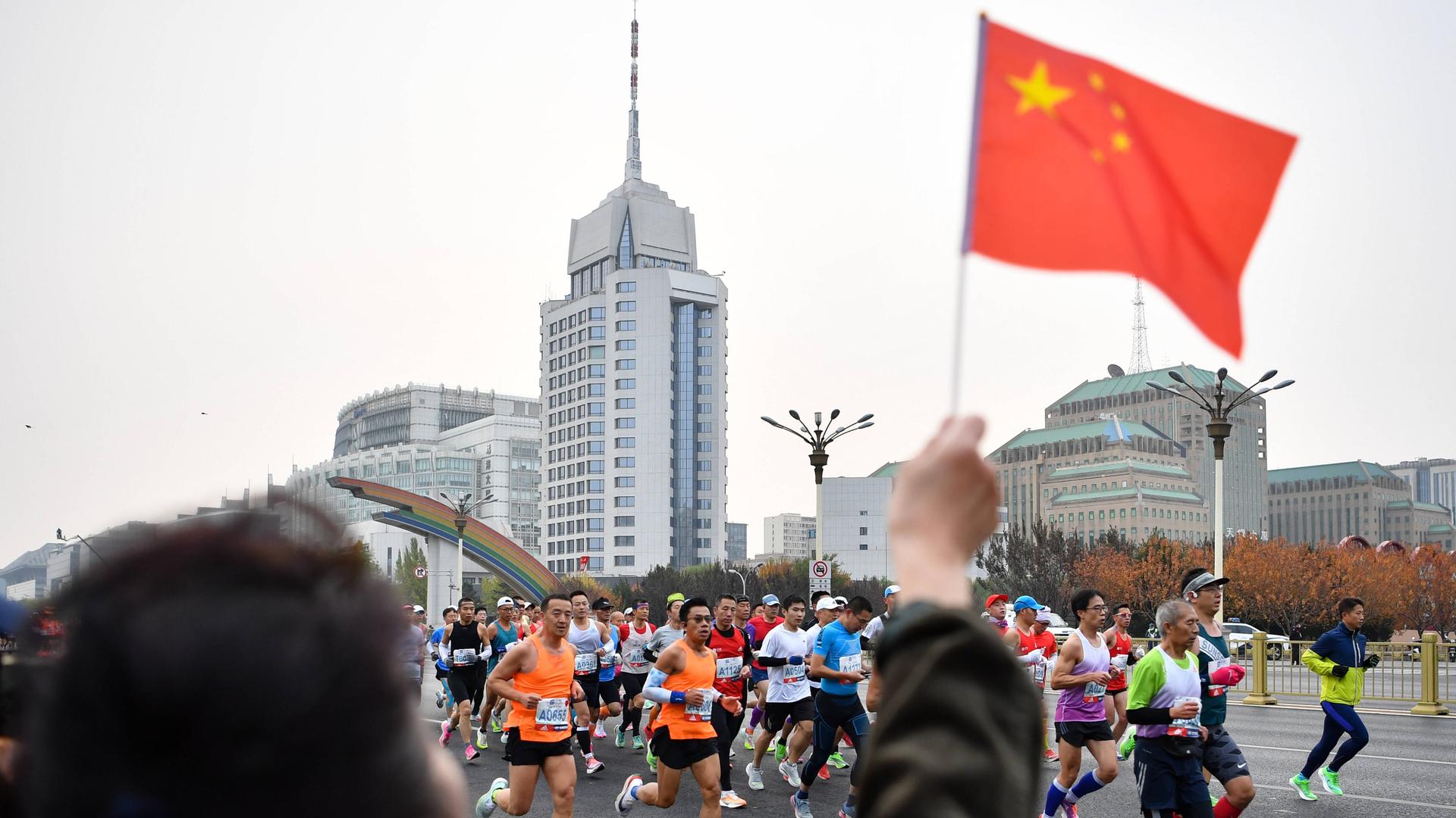 Sportlerinnen und Sportler in bunter Laufkleidung bahnen sich ihren Weg durch die chinesische Hauptstadt. Am Rand stehen Zuschauer und feuern die Teilnehmenden an. Eine Person winkt mit einer kleinen chinesischen Flagge.