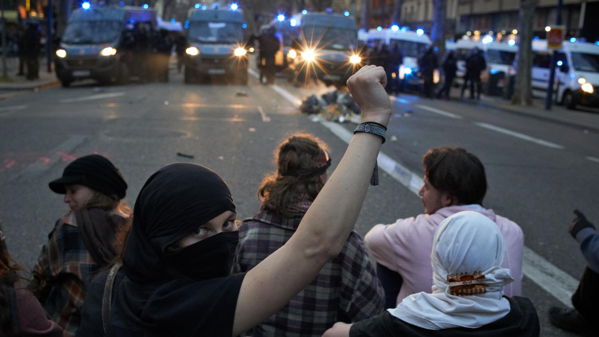 Junge Protestierende gegen die Rentenreform sitzen auf einer Straße in Toulouse. Eine verhüllte Frau reckt die rechte Faust und schaut in die Kamera. Im Hintergrund sind Polizeiwagen zu sehen.