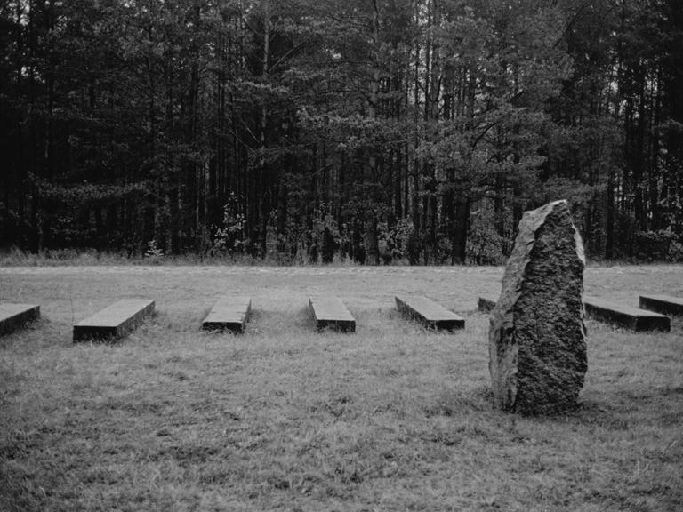 Schwarzweiß-Aufnahme einer Wiese mit Gedenkstein und Balken im Gras, die in ihrer Anordnung an Bahngleise erinnern