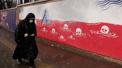 Eine in einen schwarzen Chador gehüllte Frau geht in der iranischen Hauptstadt Teheran an einem Mauergemälde entlang, das unter anderem Totenschädel zeigt. Es schneit.