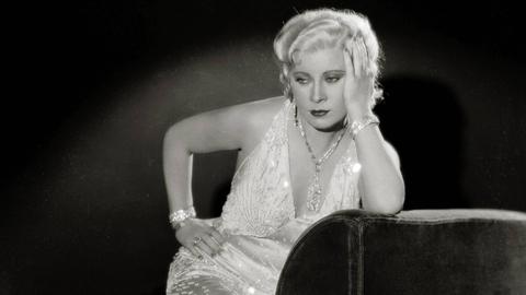 Eine historische schwarzweiss Aufnahme der Hollywood-Schauspielerin Mae West in einem glitzernden Abendkleid.