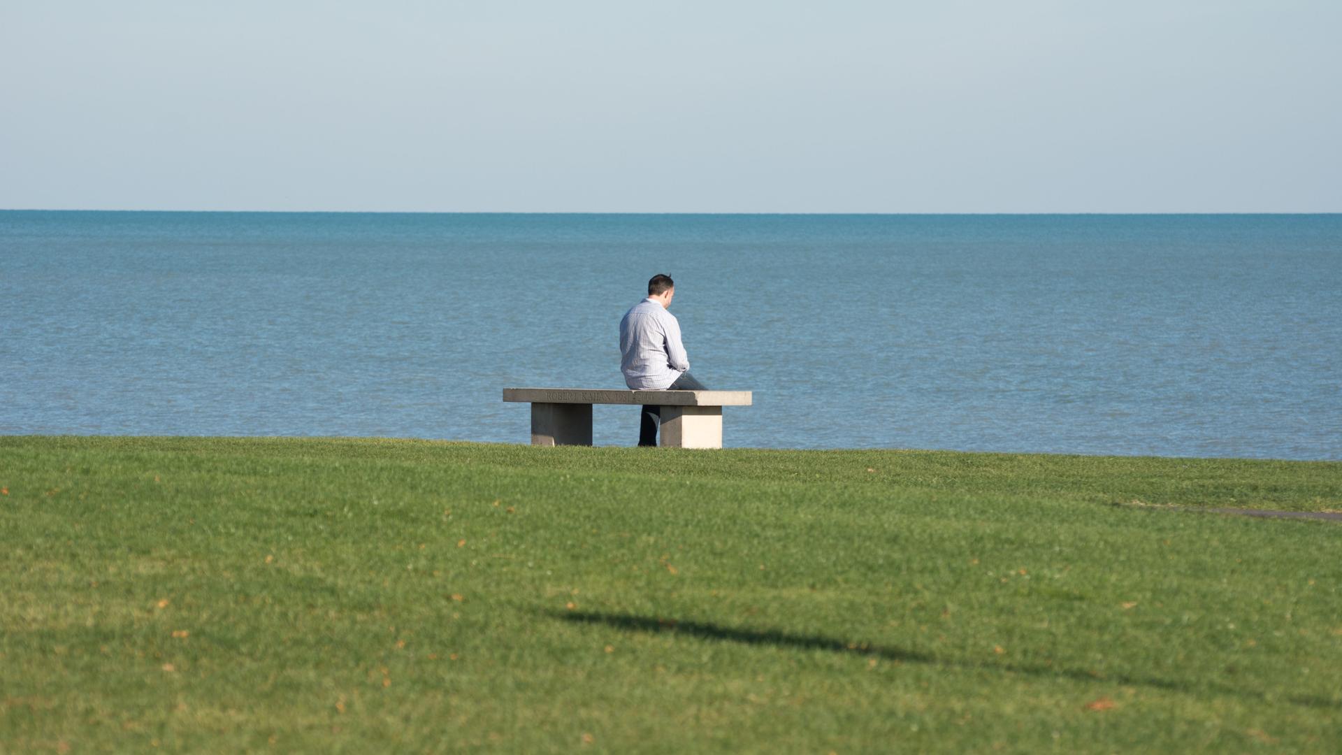 Eine Person sitzt allein auf einer Bank mit leicht gesenktem Kopf vor dem Meer. Eine Person sitzt allein auf einer Bank mit leicht gesenktem Kopf vor dem Meer.