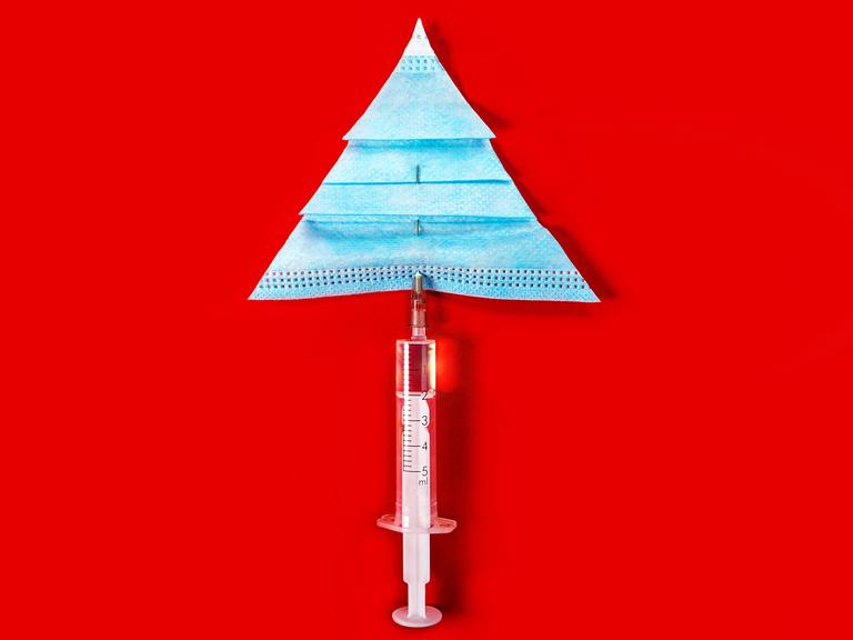 Eine Impfspritze und eine Corona-Maske, die vor rotem Hintergrund einen Tannenbaum bilden.