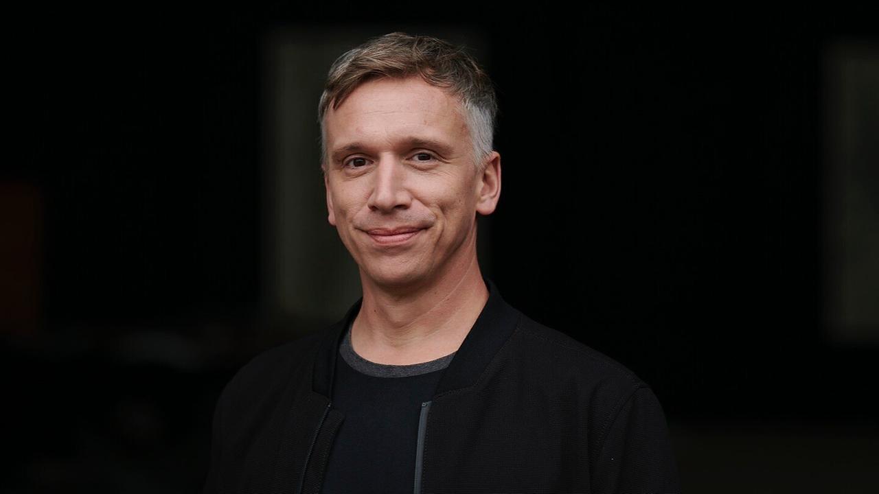 Ein Mann mit kurzen braun-grauen Haaren trägt eine schwarze dünne Jacke und lächelt in die Kamera. Der Hintergrund ist schwarz. Ein Mann mit kurzen braun-grauen Haaren trägt eine schwarze dünne Jacke und lächelt in die Kamera. Der Hintergrund ist schwarz.
