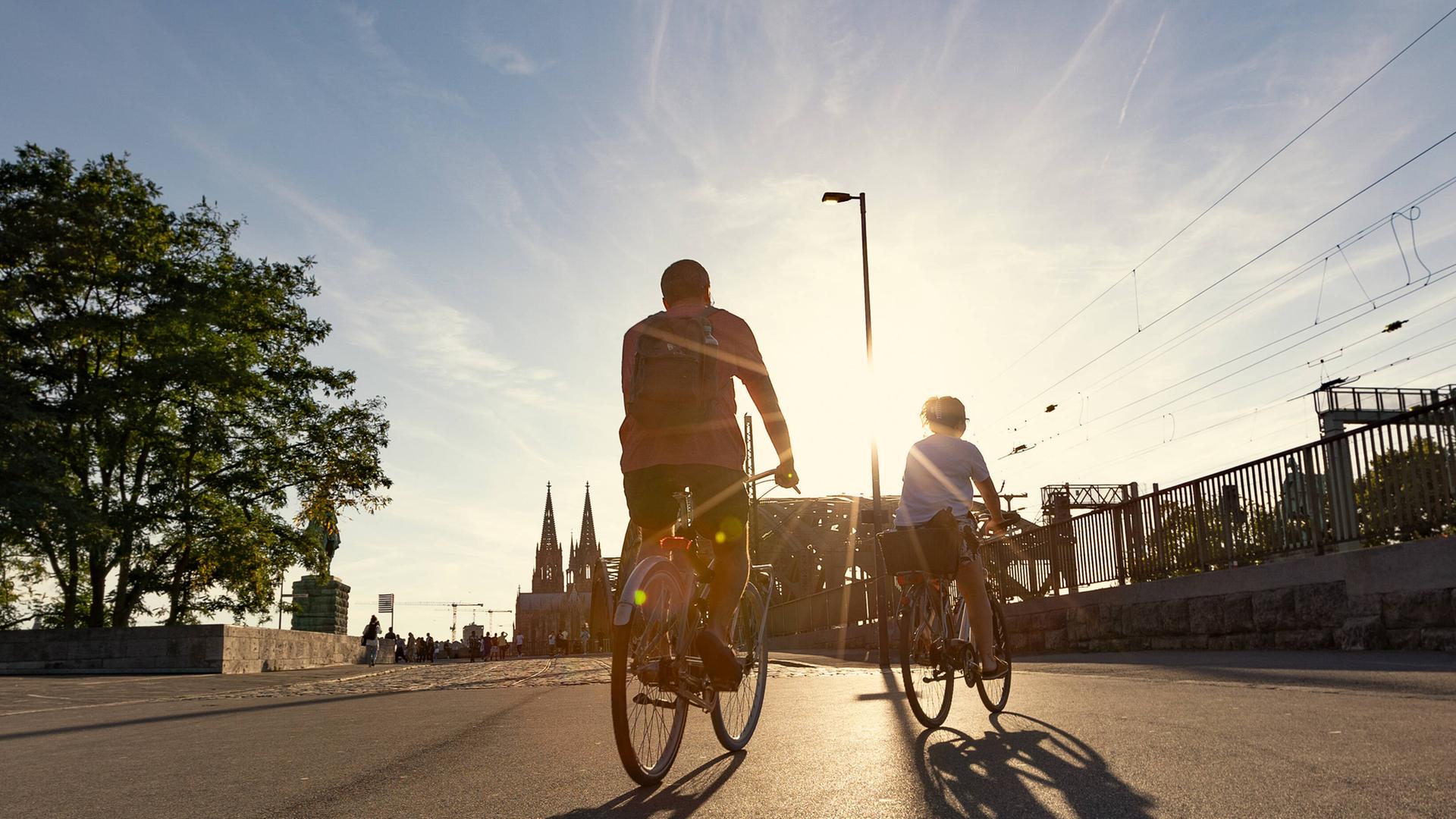 Zwei Fahrradfahrer radeln auf einer Straße in einer Stadt der Sonne entgegen.