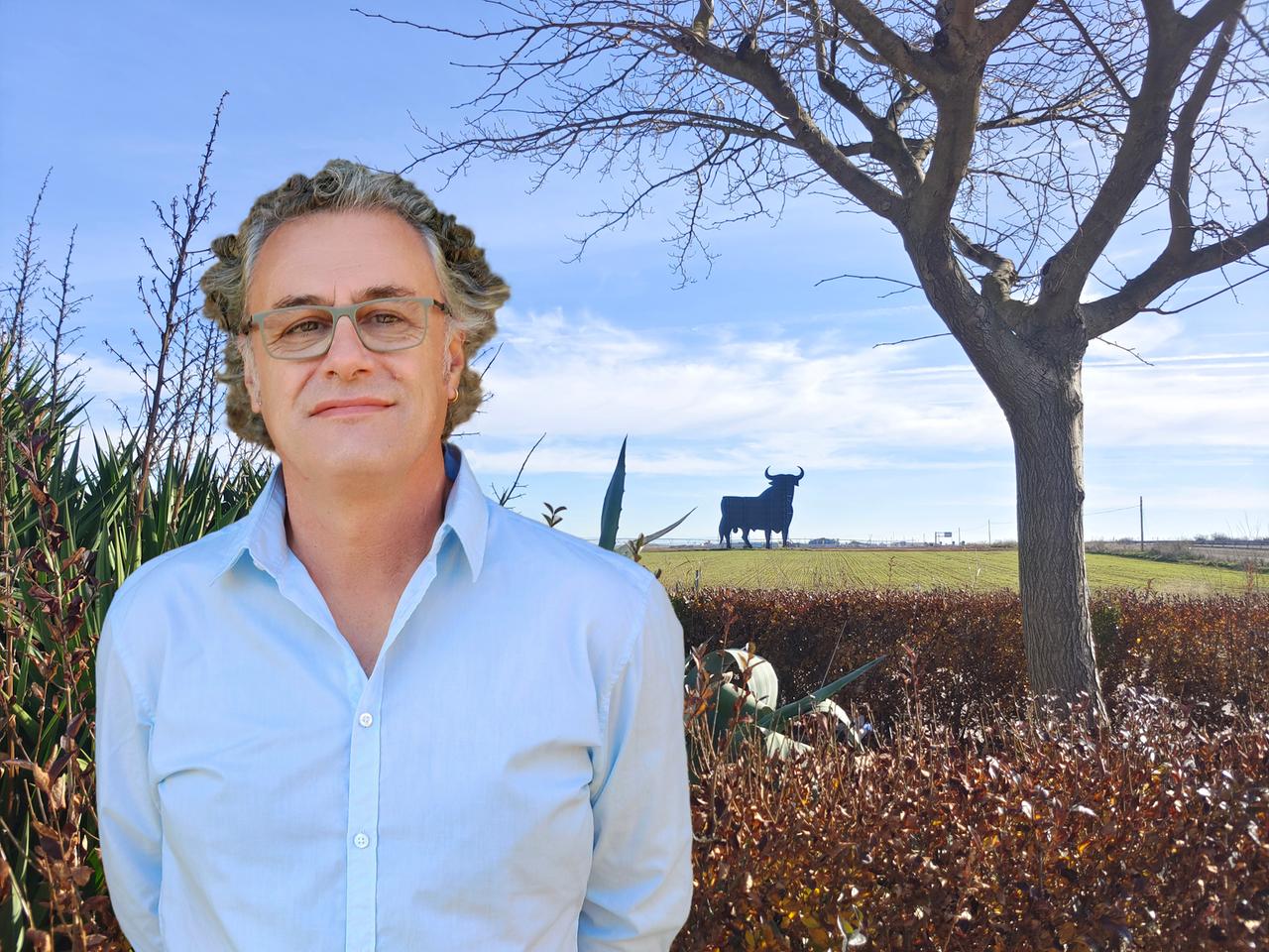 Ein Mann mit graumelierten Locken, Brille und hellblauem Hemd steht vor einem Feld auf dem am Horizont ein Stier steht.