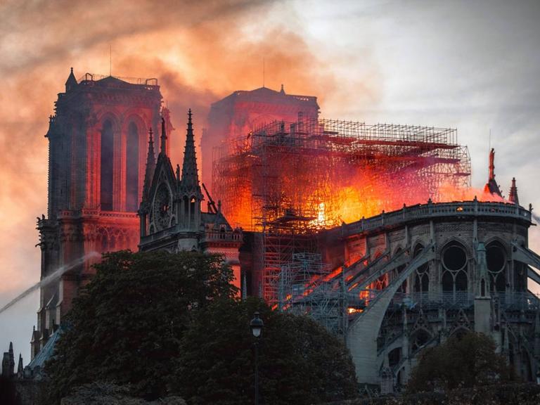 Eine Aufnahme aus dem Film "Notre-Dame in Flammen". Die bekannte Pariser Kirche brennt, die Flammen schlagen durch den Dachstuhl, am Rand wird versucht, mit Wasser zu löschen.