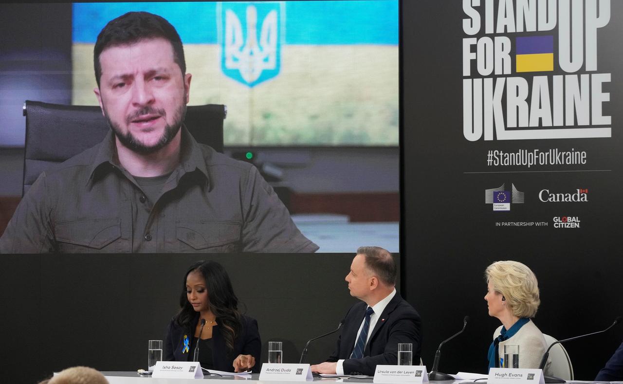 Zwei Frauen und ein Mann sitzen auf einem Podium und schauen auf eine Videoleinwand. Rechts ist ein großer Schriftzug zu sehen, der übersetzt "Steh' auf für die Ukraine" lautet. Zwei Frauen und ein Mann sitzen auf einem Podium und schauen auf eine Videoleinwand. Rechts ist ein großer Schriftzug zu sehen, der übersetzt "Steh' auf für die Ukraine" lautet.
