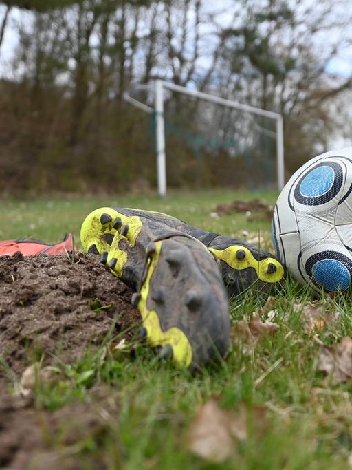 Symbolbild Fußball. Ein Spielball ist platt, ohne Luft. Fussballschuhe liegen auf einem schlechten Rasen mit Maulwurfshügeln