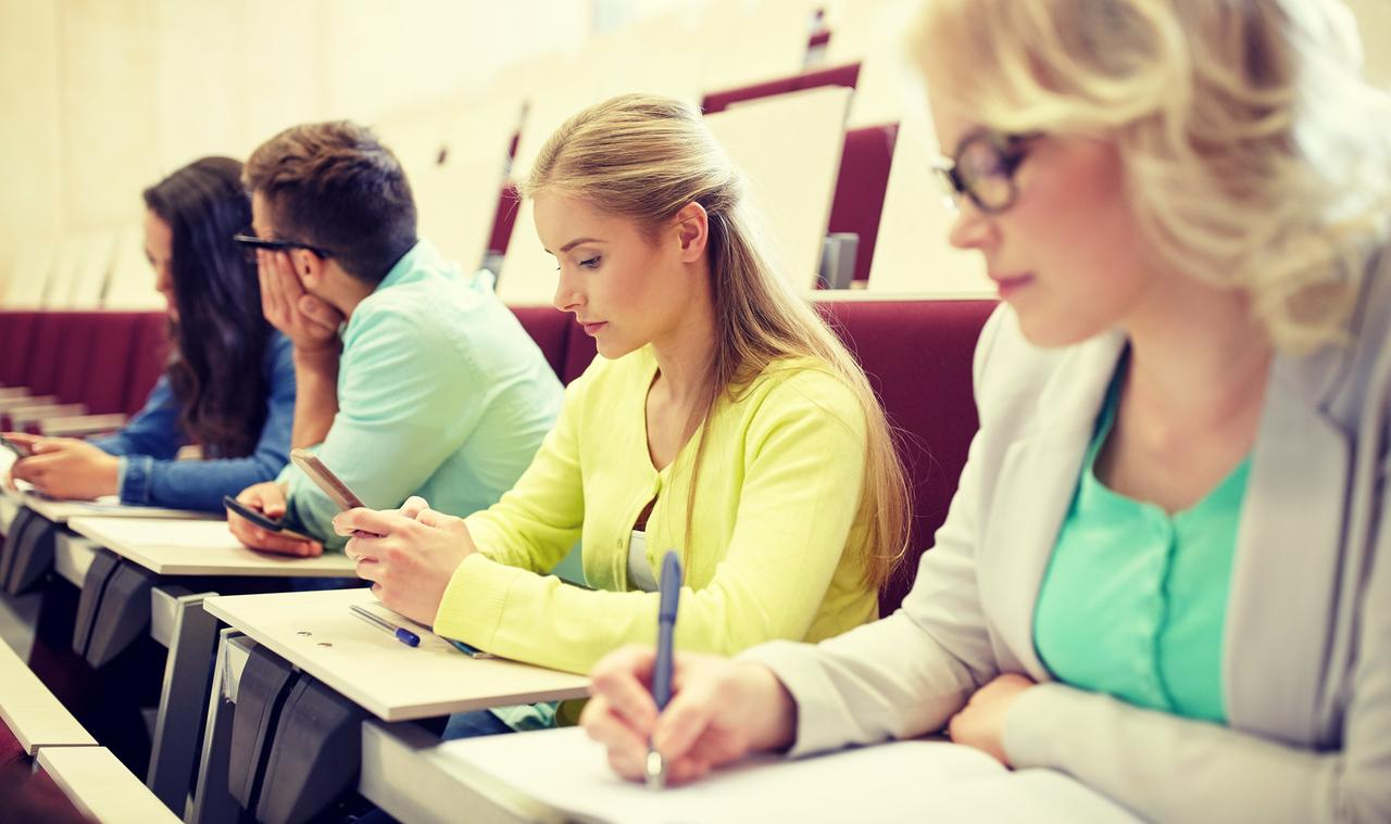 Studierende in einem Vorlesungssaale mit Notizblöcken und Smartphone