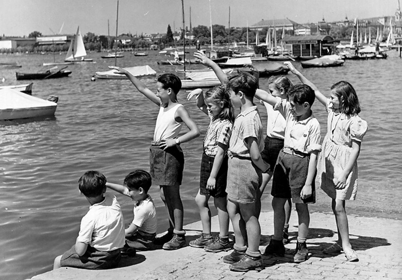 Acht Kinder stehen auf dem Schwarz.Weiß-Bild an einer Kaimauer, im Hintergrund sind Boote zu sehen, das Wasser ist ruhig, Acht Kinder stehen auf dem Schwarz.Weiß-Bild an einer Kaimauer, im Hintergrund sind Boote zu sehen, das Wasser ist ruhig,
