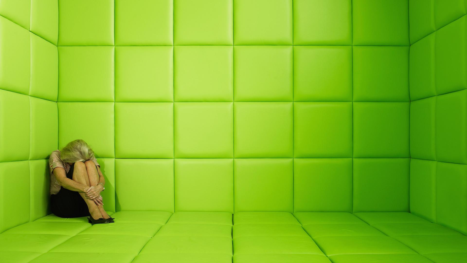 Eine Frau sitzt in der Ecke einer grün verkleideten Gummizelle und verbirgt den Kopf in ihren Armen.