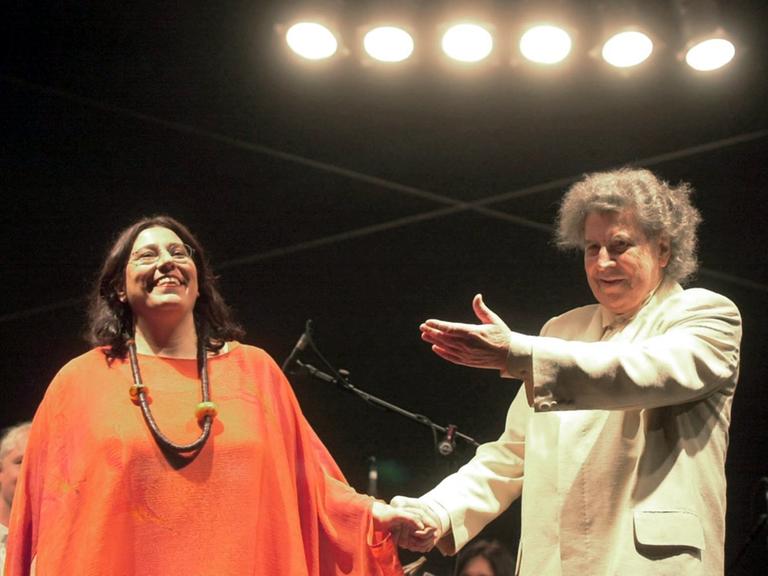Eine Frau links und ein Mann rechts stehen auf einer Bühne. Sie freuen sich. Er zeigt mit seiner Armbewegung, dass er ihr für ihren Auftritt dankt. Er ist der Komponist Mikis Theodorakis, sie die Sängerin Maria Farantouri.