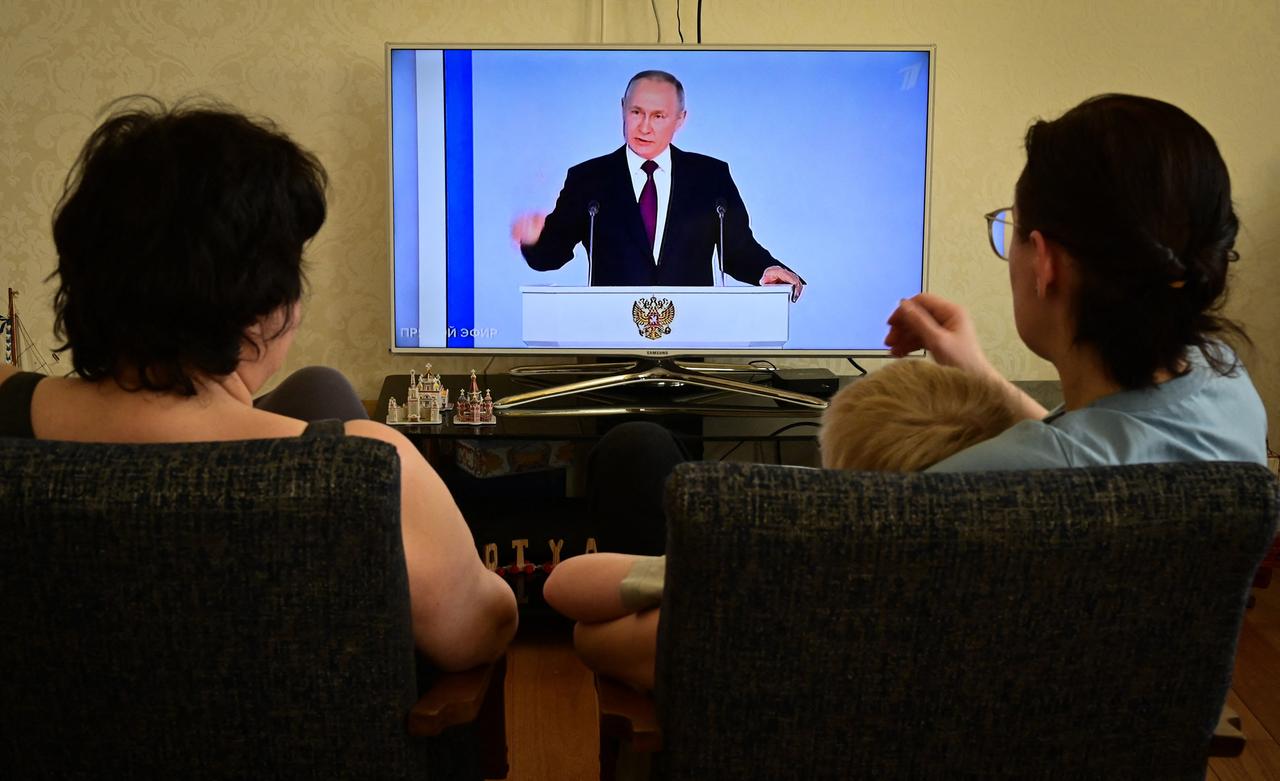 Der russische Präsident Putin spricht in einer TV-Übertragung, zwei Personen schauen zu. 
