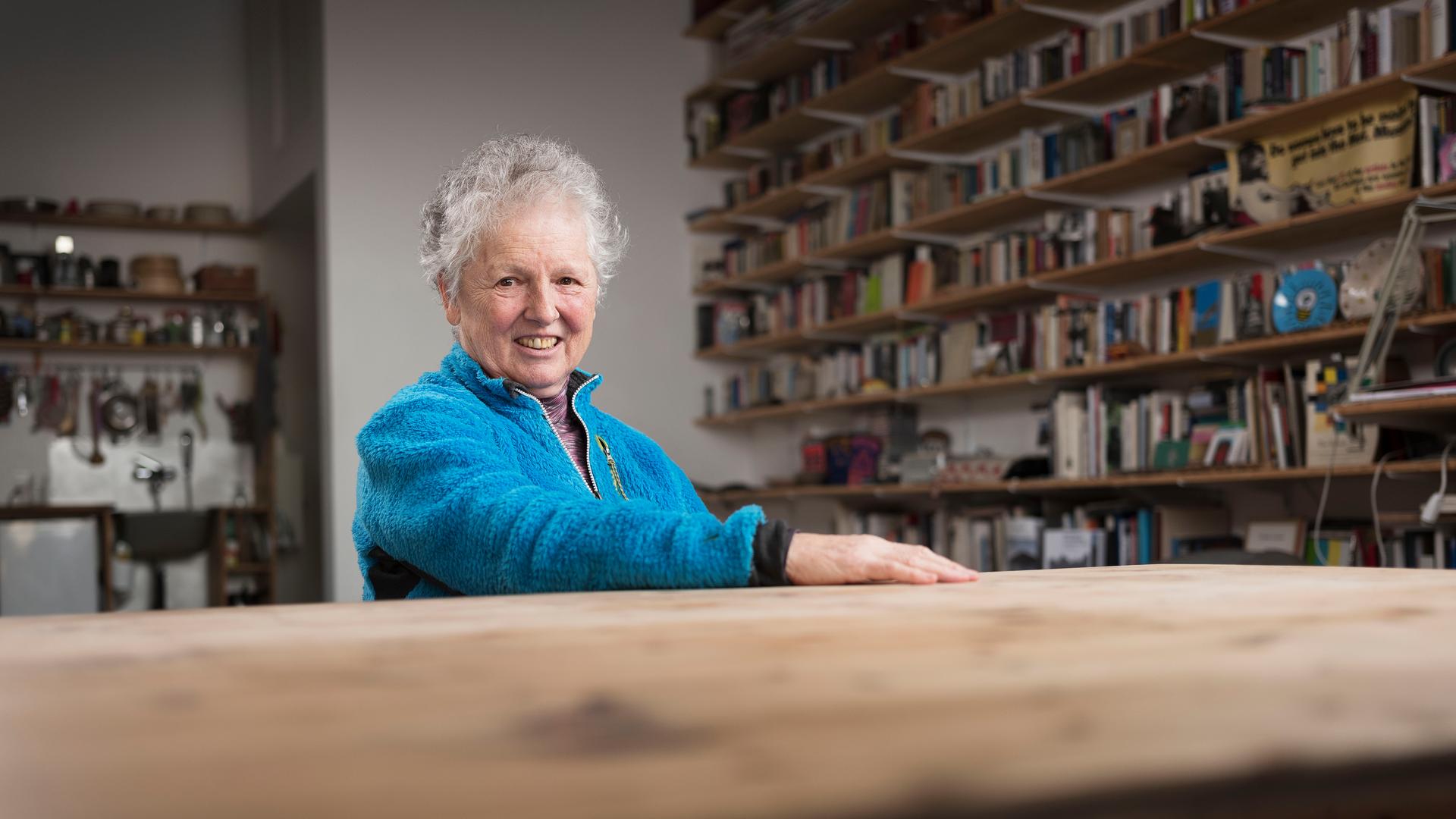 Die Künstlerin Miriam Cahn sitzt an einem Tisch und lächelt in die Kamera. Sie hat graue Haare und trägt eine blaue Jacke. Im Hintergrund sind lange Bücherregale zu sehen.