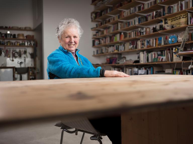 Die Künstlerin Miriam Cahn sitzt an einem Tisch und lächelt in die Kamera. Sie hat graue Haare und trägt eine blaue Jacke. Im Hintergrund sind lange Bücherregale zu sehen.