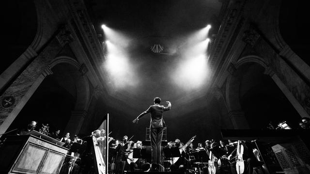 Ein Bild in schwarzweiß, das einen großen Altarraum zeigt. Von oben strahlen vier Scheinwerfer effektvoll auf ein Orchester, das dort spielt. Im Vordergrund sieht man einen hageren Mann, der die Arme hebt. Es ist der Dirigent Raphaël Pichon.