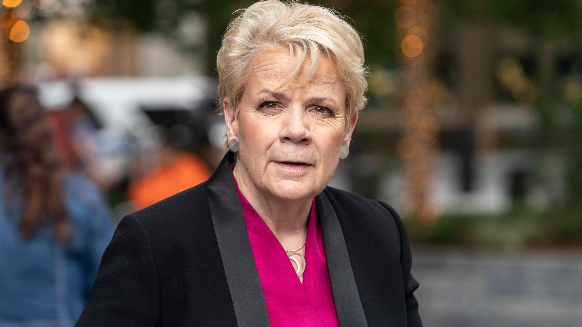 Eine ältere Frau mit kurzen blonden Haaren blickt freundlich in die Kamera. Sie trägt eine pinkfarbene Bluse und eine schwarze Anzugjacke. Hinter ihr ist nur unscharf die Umgebung zu erkennen. Eine ältere Frau mit kurzen blonden Haaren blickt freundlich in die Kamera. Sie trägt eine pinkfarbene Bluse und eine schwarze Anzugjacke. Hinter ihr ist nur unscharf die Umgebung zu erkennen.