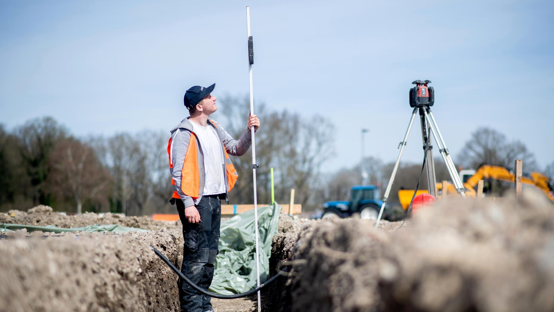 Henry Heidrich, Auszubildender in einem Bauunternehmen, steht auf einer Baustelle und misst die Tiefe eines Grabens für das Fundament mit einem Rotationslaser Henry Heidrich, Auszubildender in einem Bauunternehmen, steht auf einer Baustelle und misst die Tiefe eines Grabens für das Fundament mit einem Rotationslaser