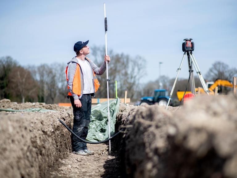 Henry Heidrich, Auszubildender in einem Bauunternehmen, steht auf einer Baustelle und misst die Tiefe eines Grabens für das Fundament mit einem Rotationslaser