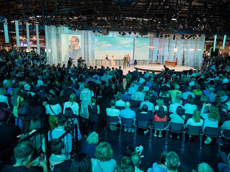 Blick aus den hinteren Reihen des Veranstaltungssaals der Republica: Viele Menschen sitzen nahe beisammen und blicken auf die perspektivisch klein wirkende Bühne. 