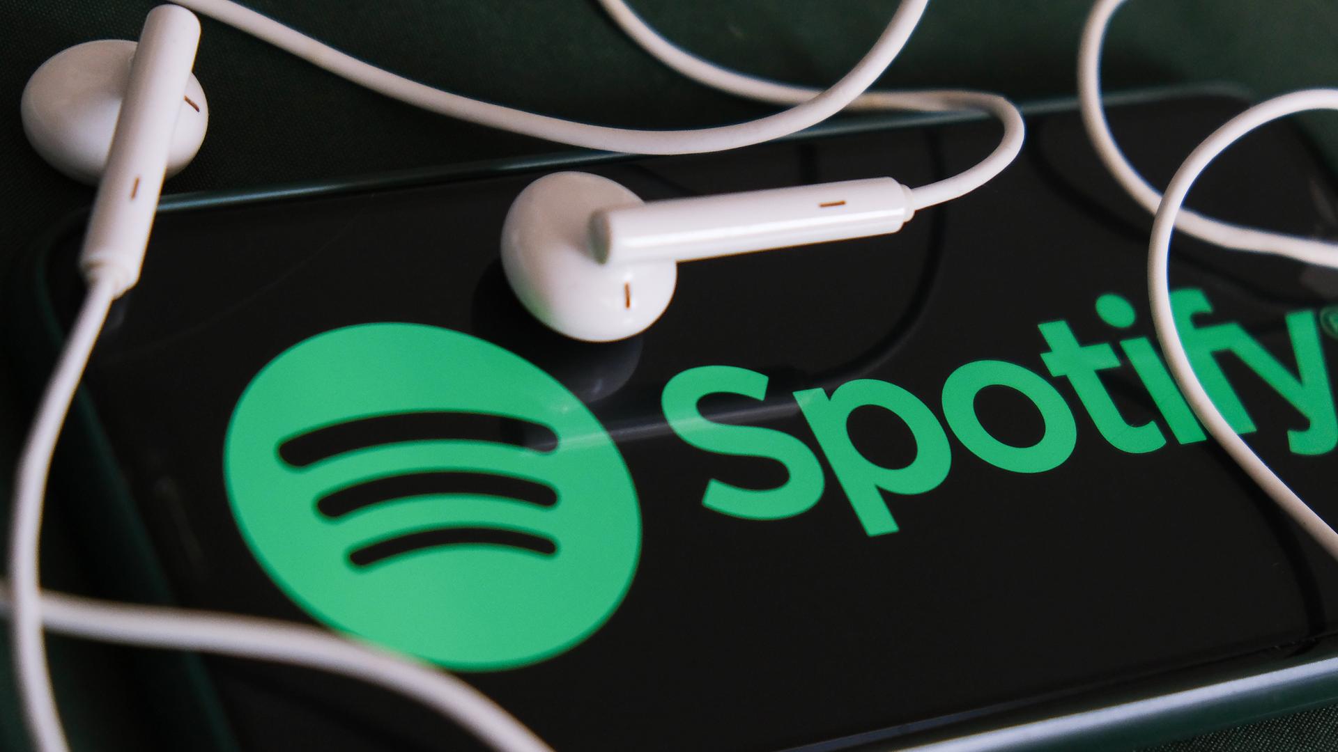 Das Foto zeigt ein Handy-Bildschirm, auf dem das Spotify in grüner Schrift angezeigt wird, und zwei Kopfhörer-Kapseln. Das Foto zeigt ein Handy-Bildschirm, auf dem das Spotify in grüner Schrift angezeigt wird, und zwei Kopfhörer-Kapseln.