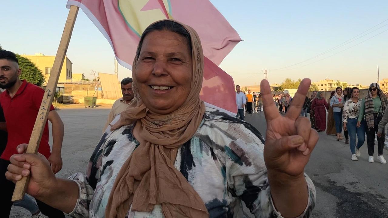 Eine ältere Frau zeigt mit zwei Fingern das Victory-Zeichen in die Kamera und lacht.