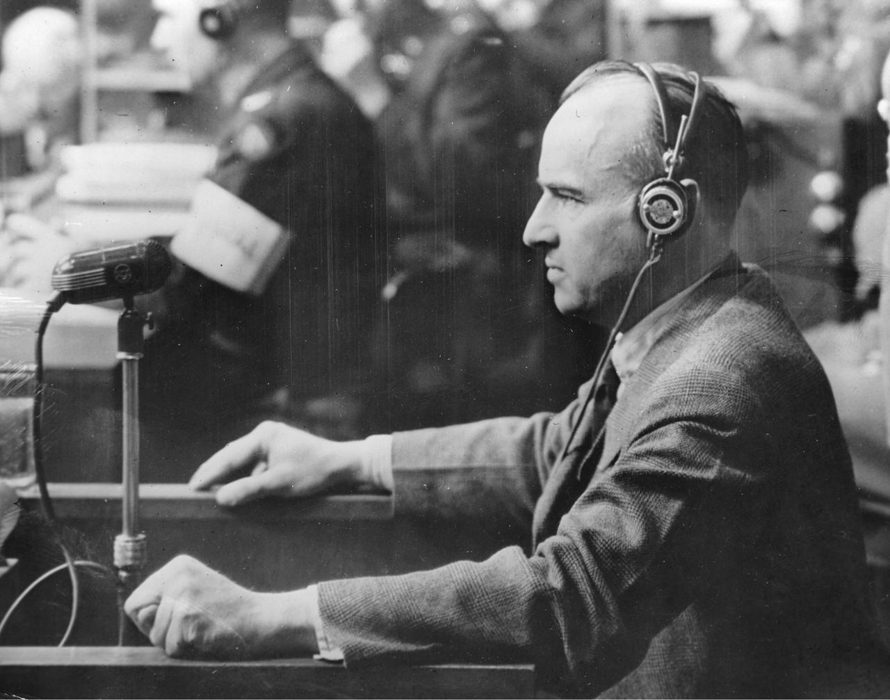 Nationalsozialist Hans Frank beim Nürnberger Prozess. Er trägt einen Kopfhörer und steht vor einem Mikrofon.