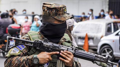 Eine Person in Militärkleidung steht mit einem Maschinengewehr im Anschlag auf einem Platz. 
