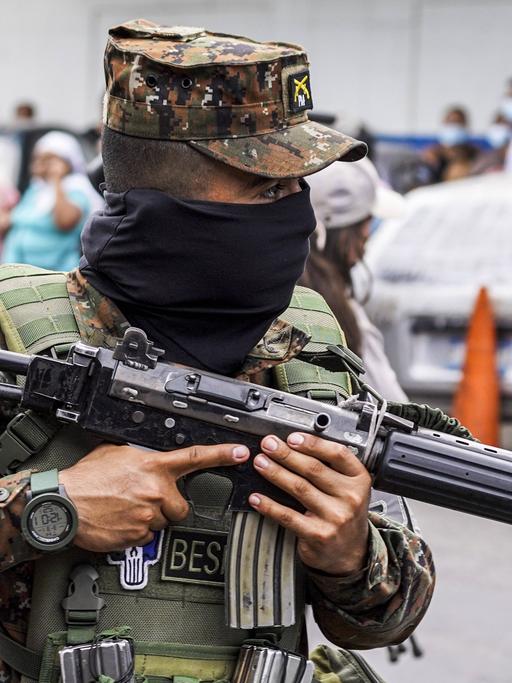 Eine Person in Militärkleidung steht mit einem Maschinengewehr im Anschlag auf einem Platz. 