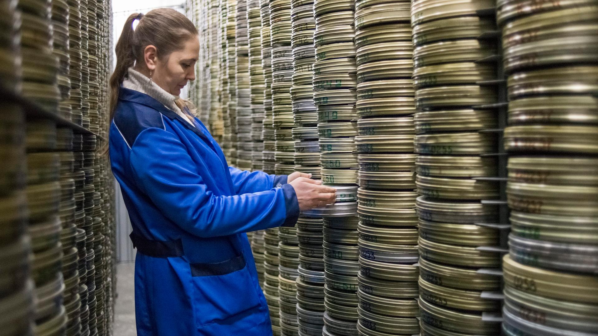 Eine Archivarin zieht in einem Raum voller gestapelter Filmrollen eine Fimrolle hervor. Eine Archivarin zieht in einem Raum voller gestapelter Filmrollen eine Fimrolle hervor.