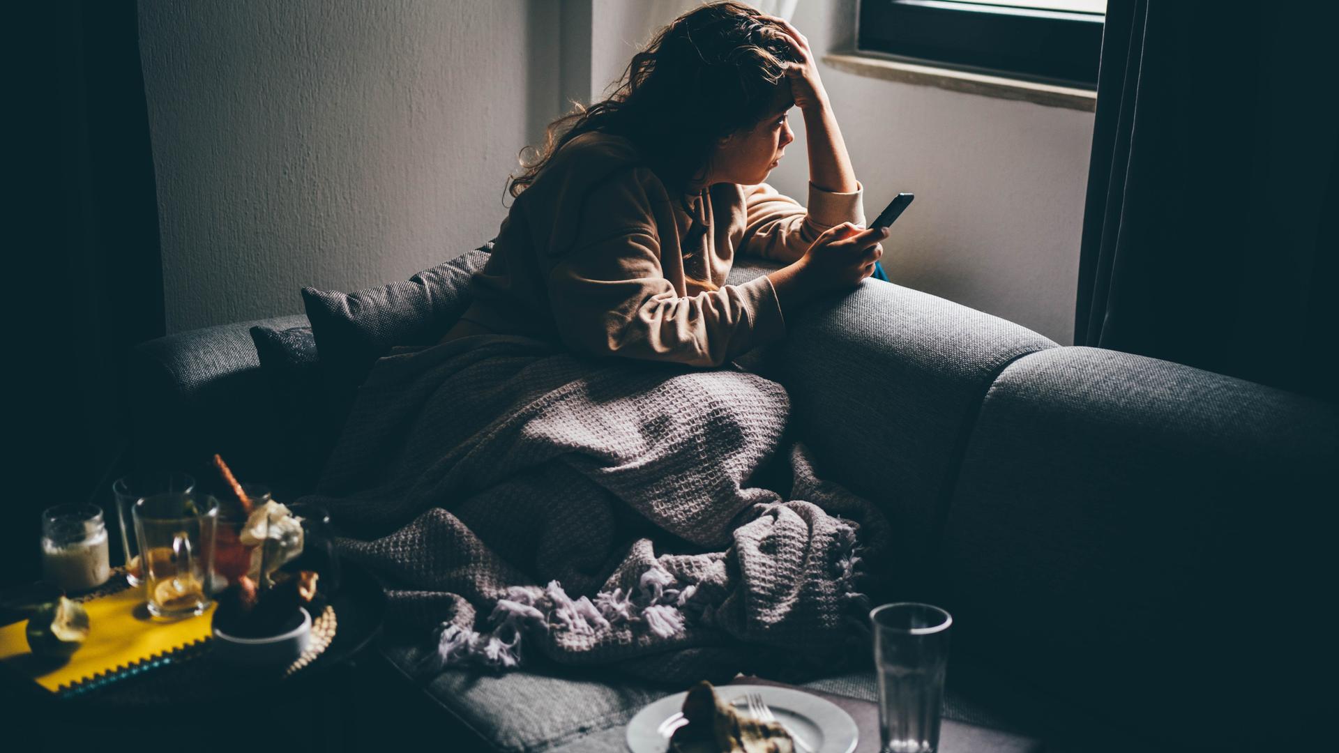 Eine junge Frau mit traurigem Gesichtsausdruck sitzt im Halbdunkel mit ihrem Smartphone in der Hand auf dem Sofa. Eine junge Frau mit traurigem Gesichtsausdruck sitzt im Halbdunkel mit ihrem Smartphone in der Hand auf dem Sofa.
