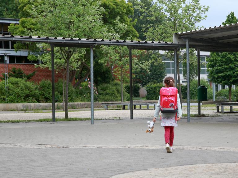 Ein Mädchen mit Schulranzen geht allein über einen Schulhof.