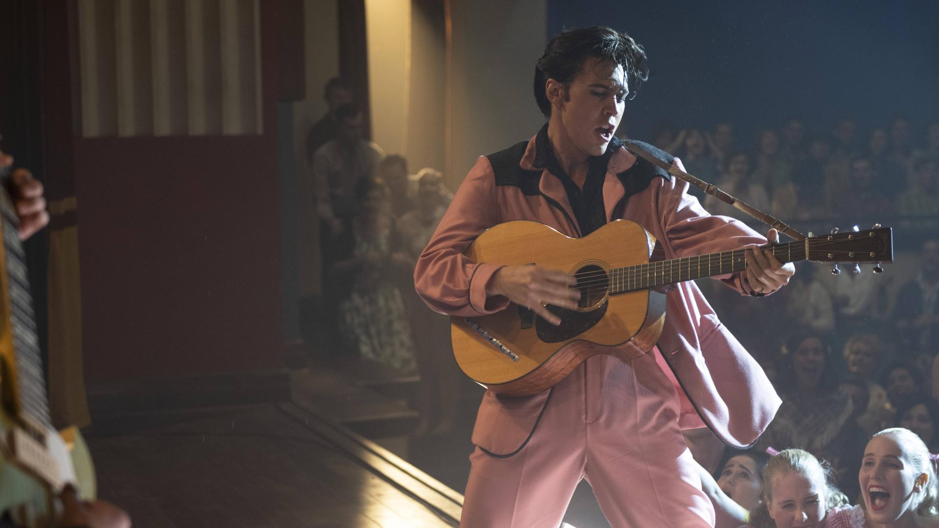 Szenenfoto aus dem Film "Elvis" von Baz Luhrmann: Schauspieler Austin Butler steht als Elvis Presley in einer Filmszene im rosa Anzug auf der Bühne, singt und spielt Gitarre. Im Hintergrund schreiende Frauen im Publikum.