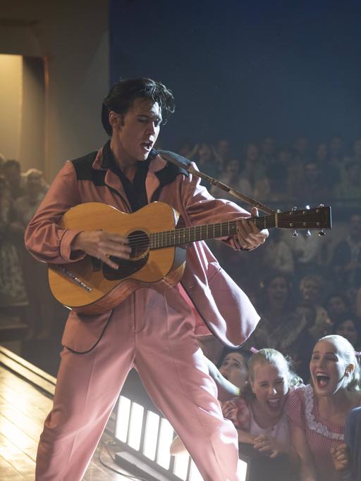 Szenenfoto aus dem Film "Elvis" von Baz Luhrmann: Schauspieler Austin Butler steht als Elvis Presley in einer Filmszene im rosa Anzug auf der Bühne, singt und spielt Gitarre. Im Hintergrund schreiende Frauen im Publikum.