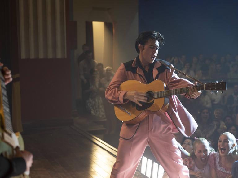 Szenenfoto aus dem Film "Elvis" von Baz Luhrmann: Schauspieler Austin Butler steht als Elvis Presley in einer Filmszene im rosa Anzug auf der Bühne, singt und spielt Gitarre. Im Hintergrund schreiende Frauen im Publikum.