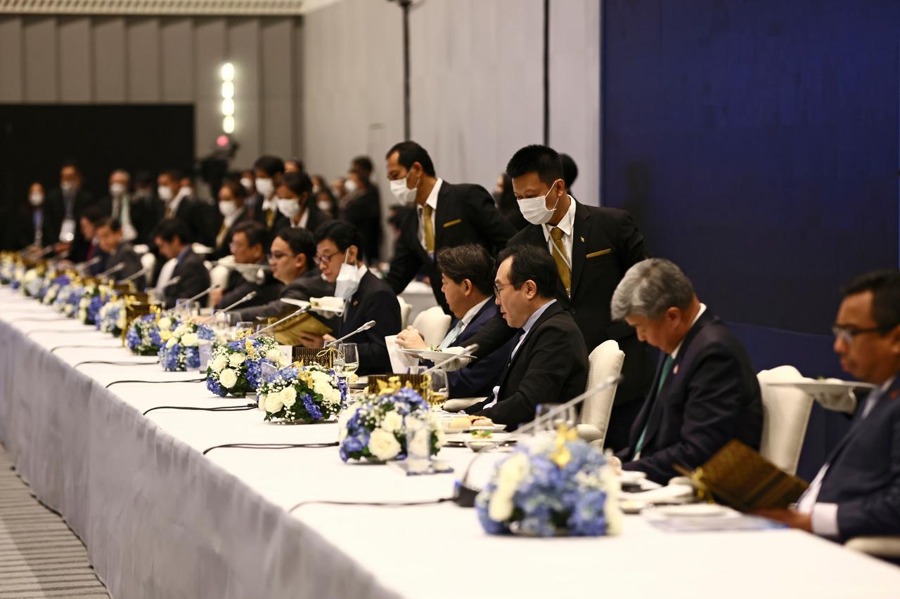 Die Teilnehmer sitzen an einem langen Tisch mit Blumengebinden. Bedienungen reichen ihnen von hinten das Essen. 