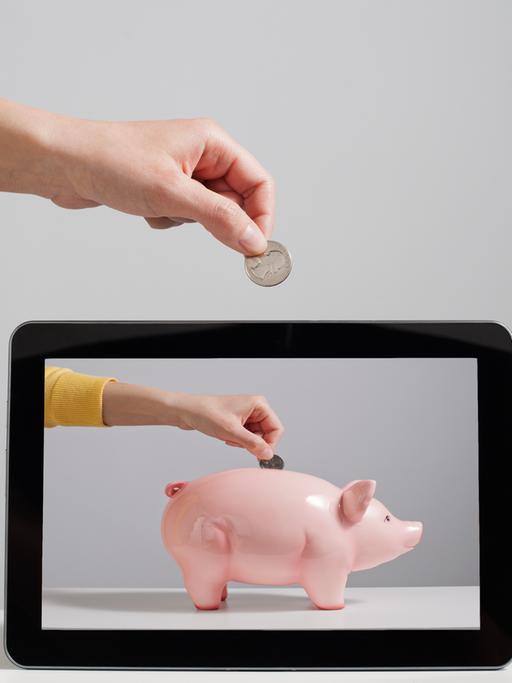 Eine Hand wirft Geld in ein Sparschwein, welches nur noch auf dem Bildschirm auftauscht.