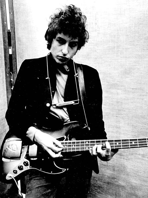 Bob Dylan in einem Studion in New York an einer Fender Jazz Bassgitarre, um seinen Hals hängt eine Mundharmonika.