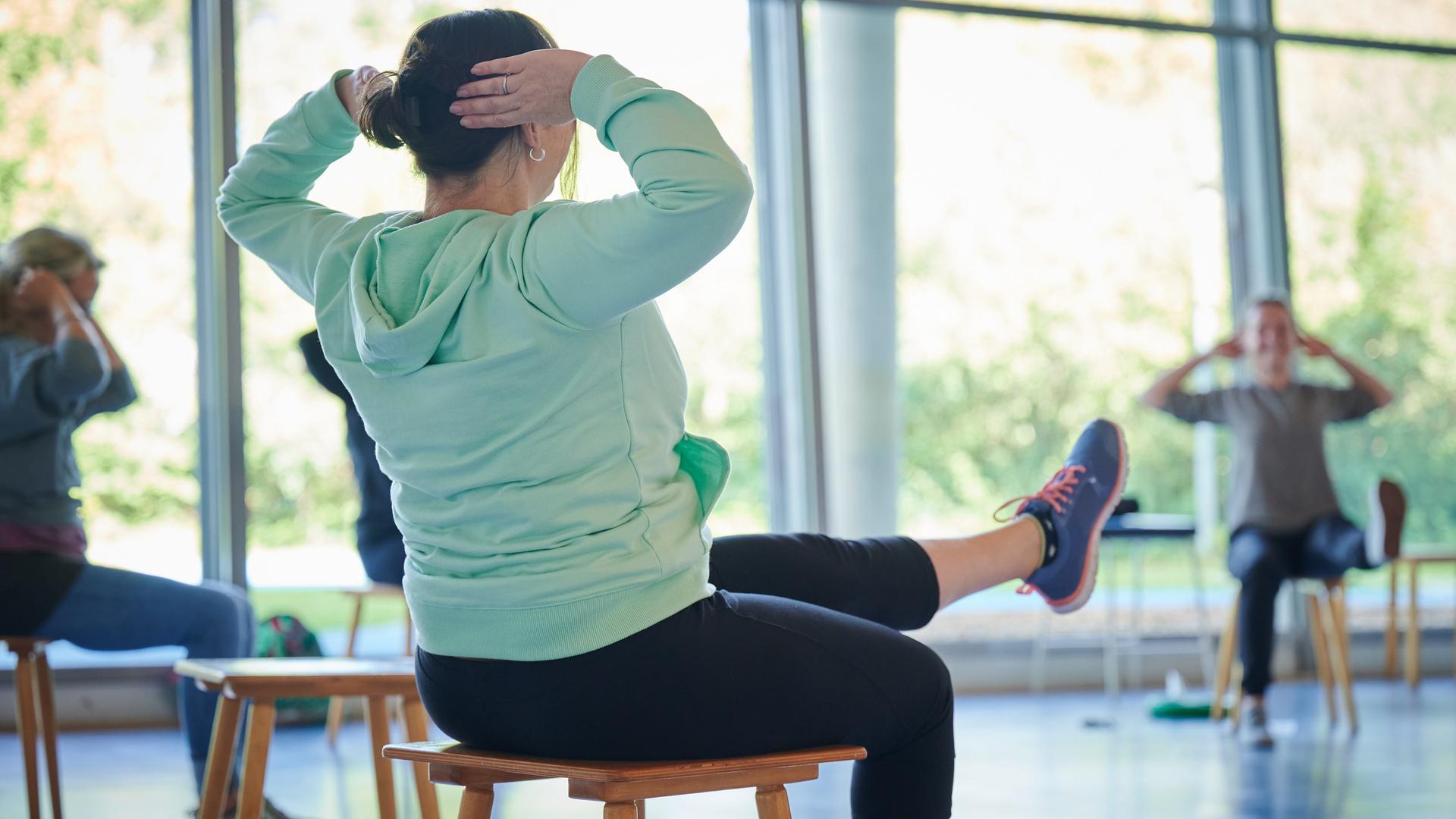 Patienten in Sportkleidung sitzen in einem großen, hellen Raum mit Abstand voneinander auf Hockern und führen eine Übung mit nach vorne ausgestrecktem Bein und hinter dem Kopf gehaltenen Armen durch.