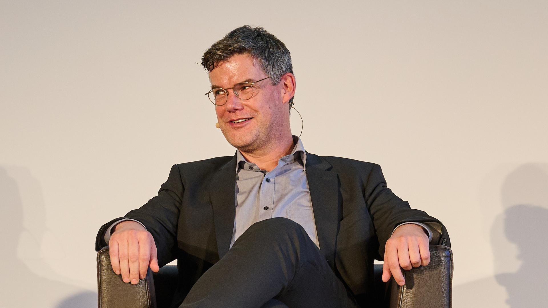 Ralph Bollmann sitzt in einem Ledersessel auf einer Bühne und spricht über seine Merkel-Biografie. Ralph Bollmann sitzt in einem Ledersessel auf einer Bühne und spricht über seine Merkel-Biografie.