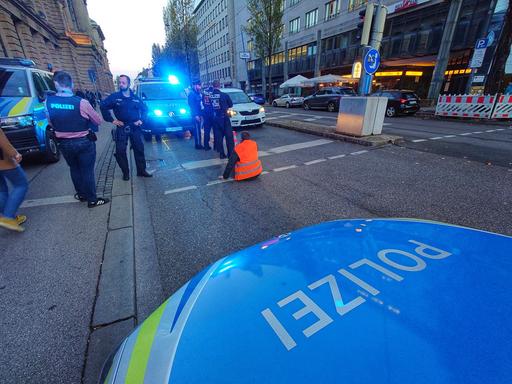 Ein Klimaaktivist hat sich am 7. November auf einer Straße in München an den Asphalt geklebt. Er träge eine rote Weste, die leuchtet. Um ihn herum stehen mit etwas Abstand mehrere Polizeiwagen und Polizisten, teilweise ist das Blaulicht an.