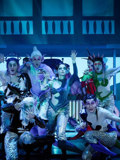 Sieben queere Fantasy-Figuren von Meerjungfrauen und -männern auf der Bühne, umrahmt von Musikern.