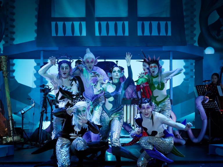 Sieben queere Fantasy-Figuren von Meerjungfrauen und -männern auf der Bühne, umrahmt von Musikern.
