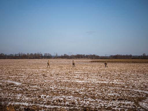 Drei ukrainische Soldaten gehen durch einen Acker, in Bildhintergrund sind weitere landwirtschaftliche Nutzfelder zu erkennen.