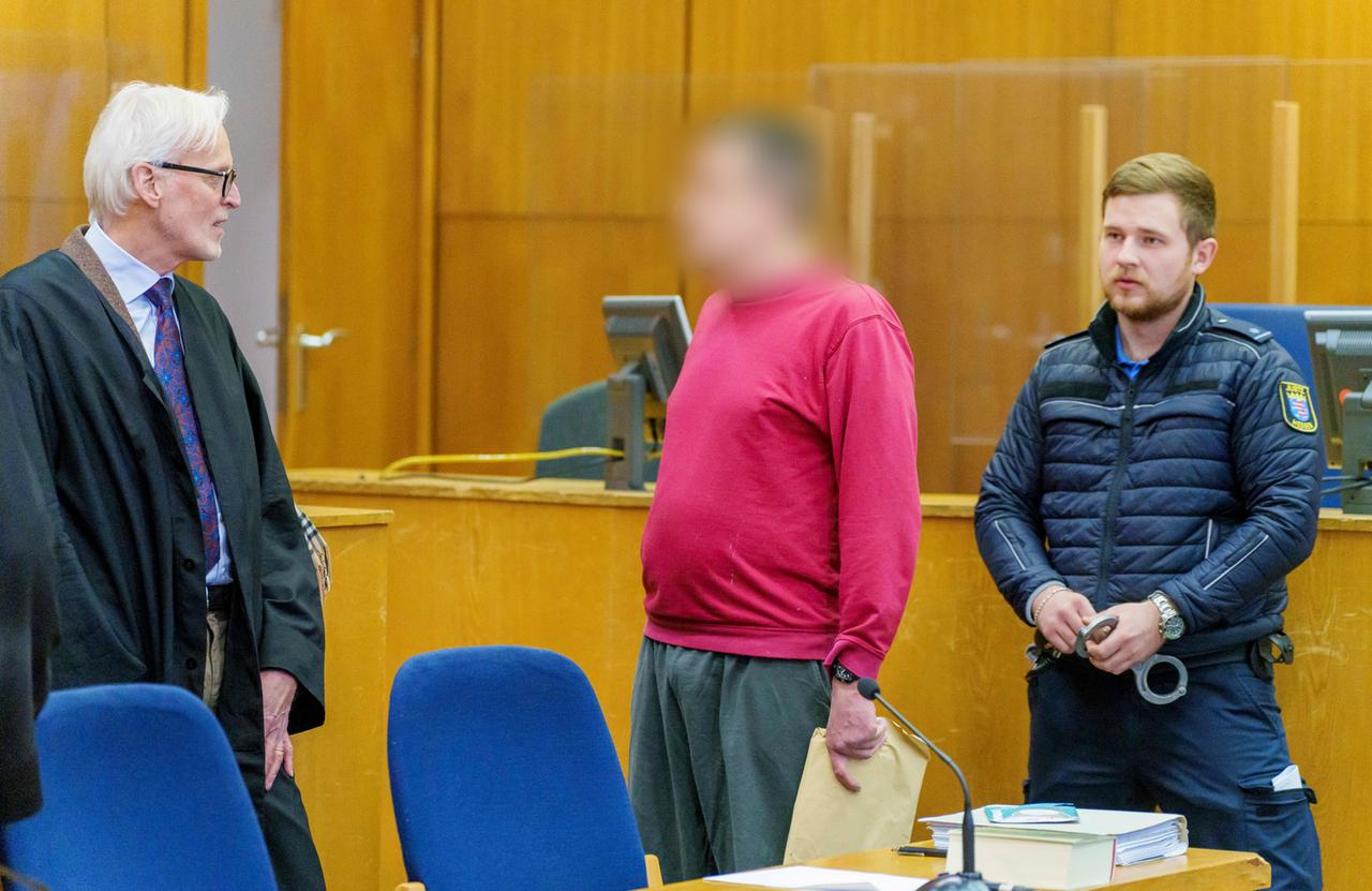 Ein Mann mit einem verpixelten Gesicht steht in einem Gerichtssaal, neben ihm ein Strafverteidiger und ein Polizist.