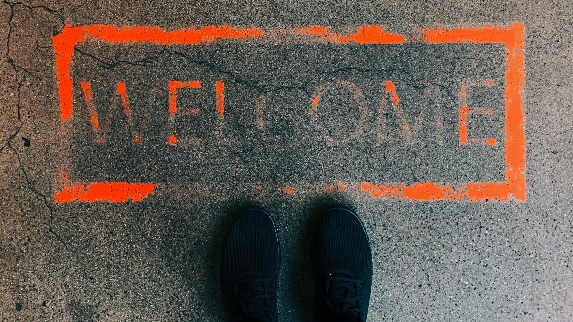 Ein orangefarbenes Rechteck auf dem Boden. Darin das Wort "Welcome". Die Farbe ist größtenteils bereits verblasst. Davor, im unteren Bildbereich: zwei Schuhspitzen.