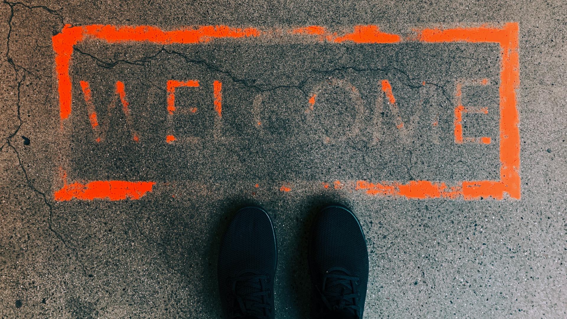 Ein orangefarbenes Rechteck auf dem Boden. Darin das Wort "Welcome". Die Farbe ist größtenteils bereits verblasst. Davor, im unteren Bildbereich: zwei Schuhspitzen. Ein orangefarbenes Rechteck auf dem Boden. Darin das Wort "Welcome". Die Farbe ist größtenteils bereits verblasst. Davor, im unteren Bildbereich: zwei Schuhspitzen.