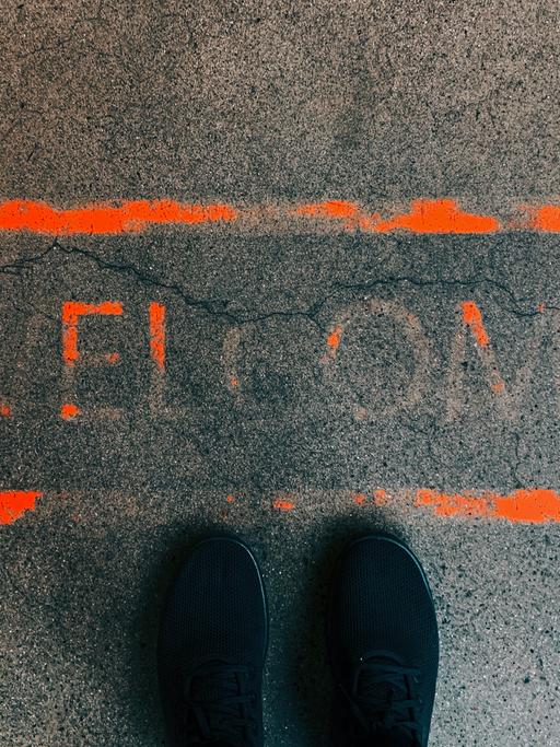 Ein orangefarbenes Rechteck auf dem Boden. Darin das Wort "Welcome". Die Farbe ist größtenteils bereits verblasst. Davor, im unteren Bildbereich: zwei Schuhspitzen.